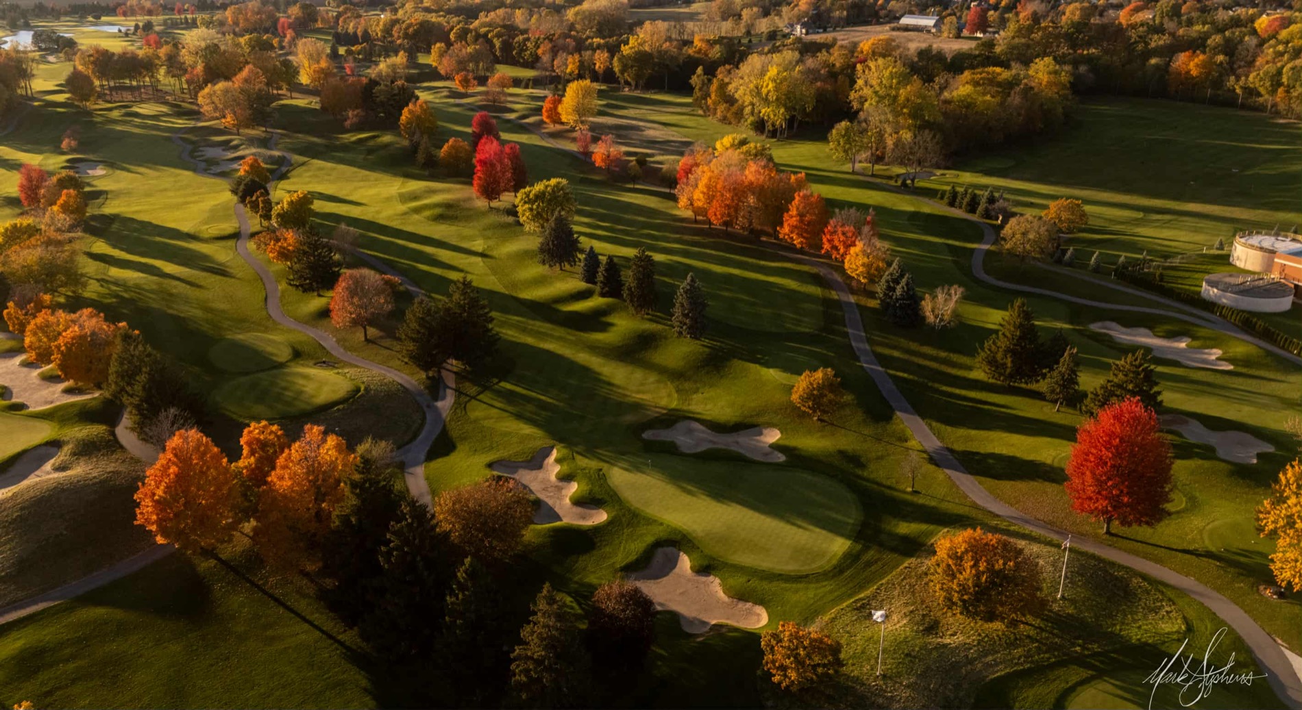 Aerial of a parkland course at last light, low sun casting long shadows down fairways studded with autumn colour