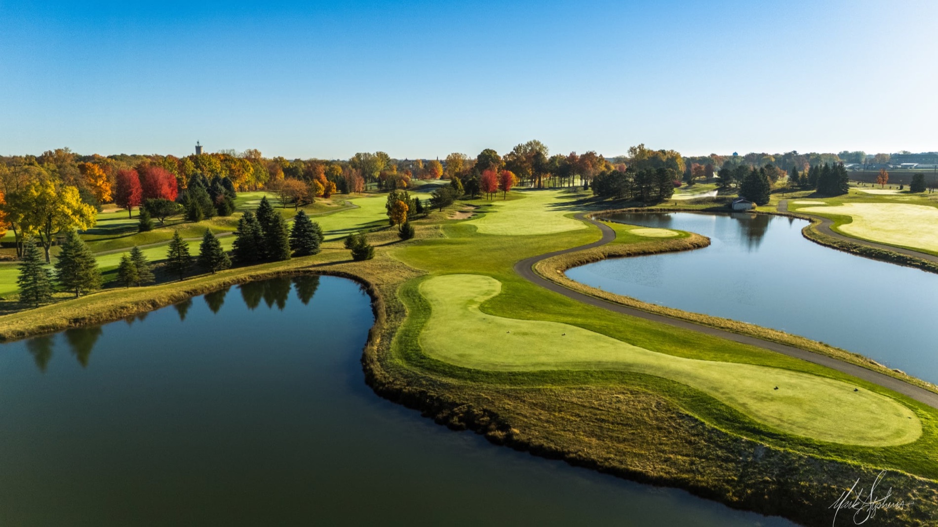 Aerial of a fairway peninsula with still water down both sides mirroring the autumn treeline under a clear blue sky