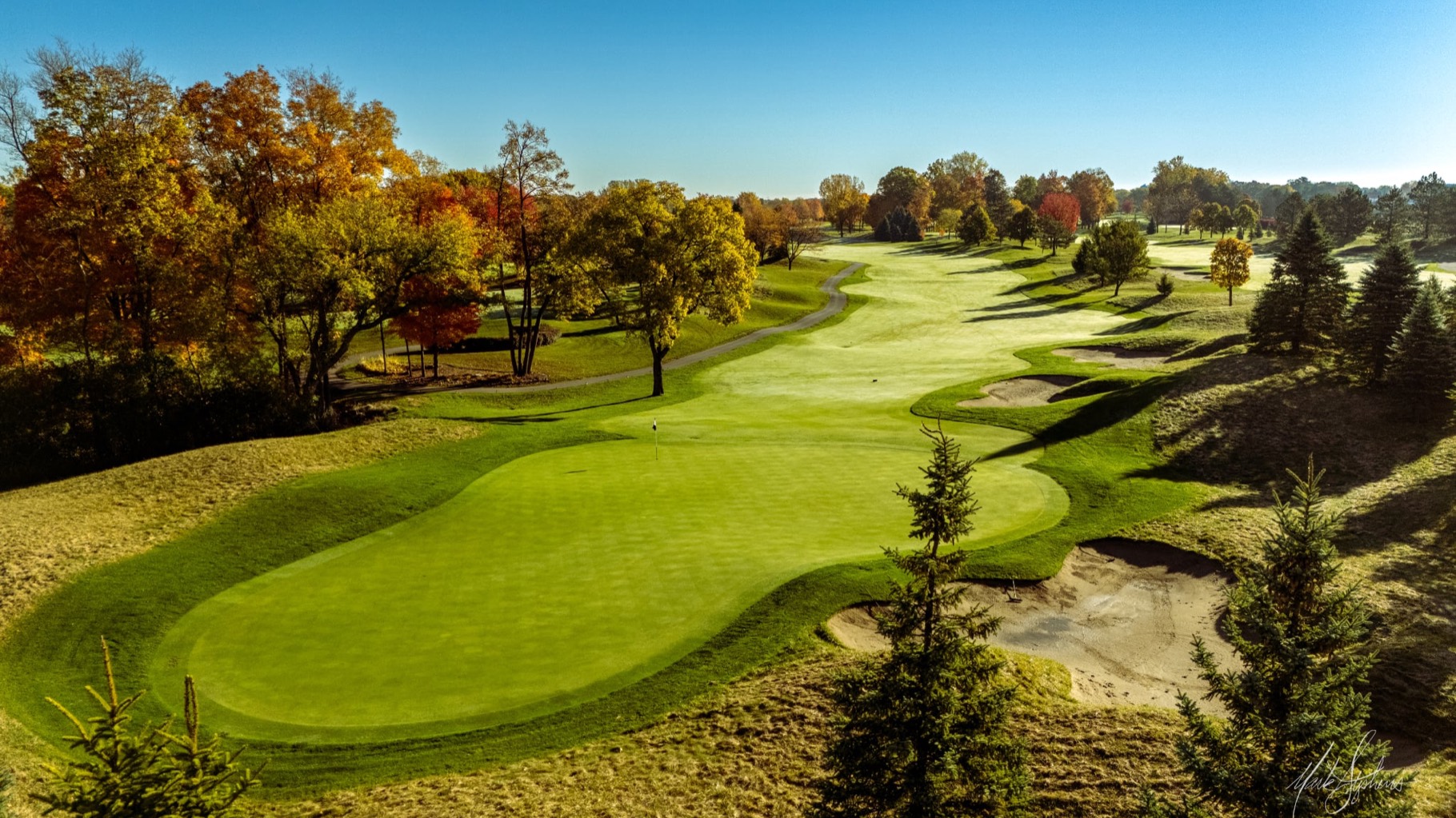 Ground-level view down a manicured approach to a green, framed by autumn hardwoods and evergreens