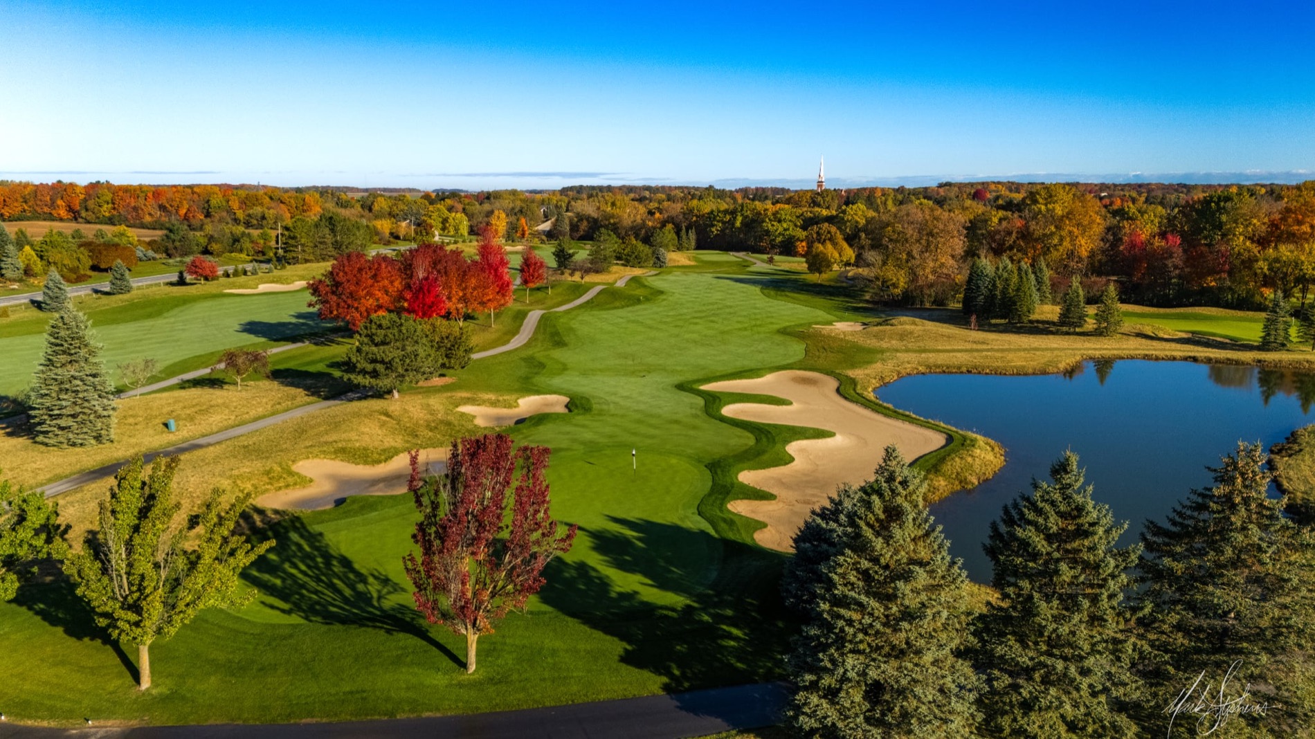 Aerial of a parkland green complex beside a pond, red maples in the foreground and a distant church spire on the horizon