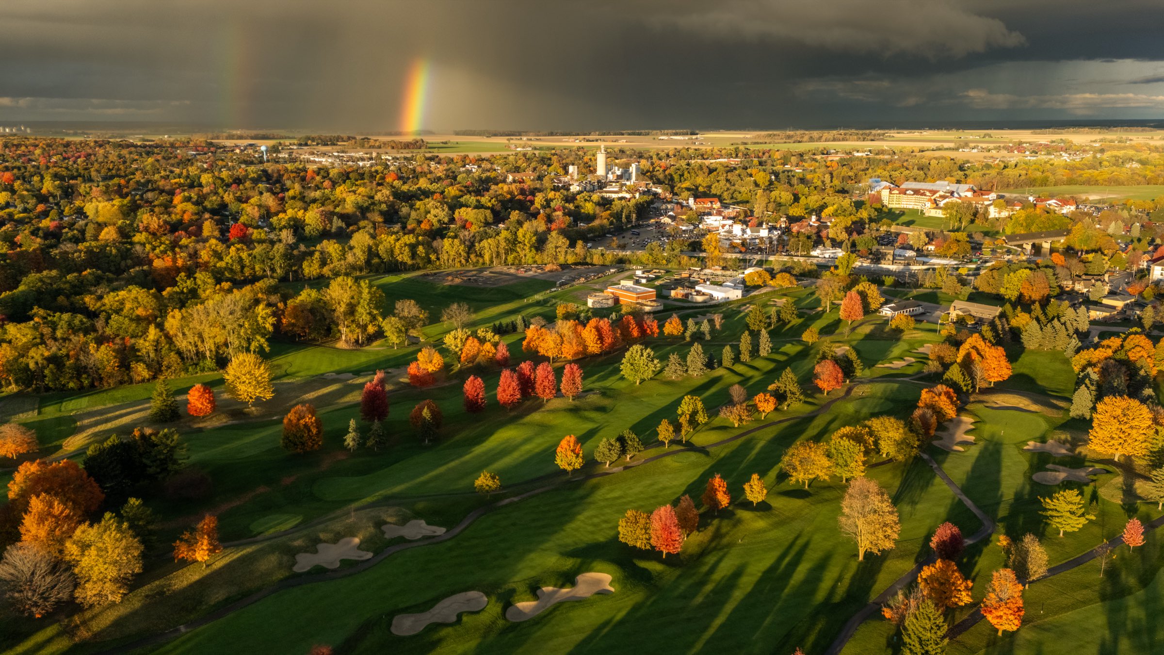Storm-light aerial of the golf course and the town beyond, a double rainbow arcing over the far autumn treeline